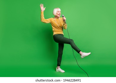 Full Size Photo Of Optimistic Nice Blond Old Lady Sing In Mic Dance Wear Yellow Cloth Trousers Sneakers Isolated On Green Background