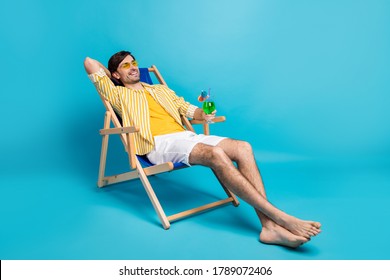 Full size photo positive guy enjoy rest relax exotic resort sun bathing hold glass cocktail sit deckchair wear white yellow striped shirt shorts barefoot isolated blue color background - Shutterstock ID 1789072406
