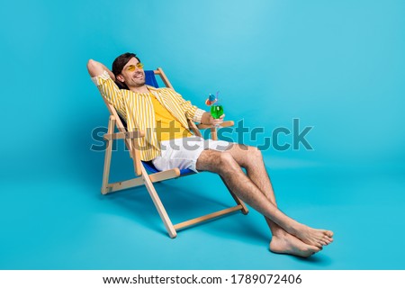 Full size photo positive guy enjoy rest relax exotic resort sun bathing hold glass cocktail sit deckchair wear white yellow striped shirt shorts barefoot isolated blue color background