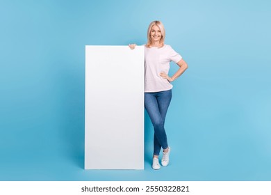 Full size photo of positive pensioner woman white t-shirt jeans sneakers stand near panel empty space isolated on blue color background - Powered by Shutterstock