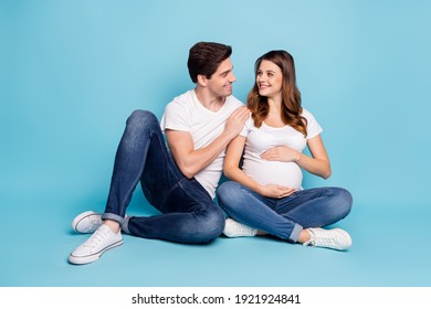 Full size photo of positive spouse look each other sit on floor touch stomach isolated on blue color background