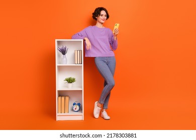 Full Size Photo Of Pretty Nice Lady Use Gadget Telephone Stand Near Book Shelf Isolated On Orange Color Background