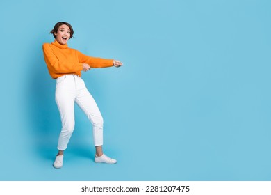 Full size photo of pretty young girl pulling rope string excited empty space dressed stylish orange look isolated on blue color background - Shutterstock ID 2281207475