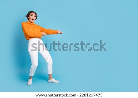 Full size photo of pretty young girl pulling rope string excited empty space dressed stylish orange look isolated on blue color background