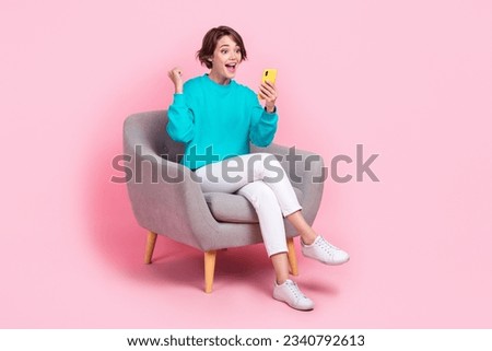 Full size photo of pretty young girl sit armchair raise fist hold telephone dressed stylish blue outfit isolated on pink color background