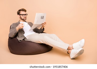 Full size photo of serious focused young man sit chair read paper drink coffee isolated on beige color background - Powered by Shutterstock