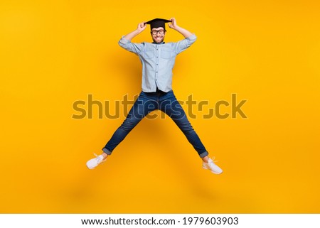 Full size photo of young man happy positive smile jump hands touch mortarboard student isolated over yellow color background