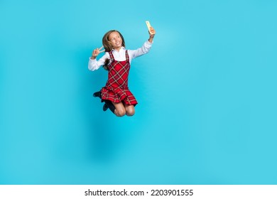 Full size portrait of small beautiful schoolgirl jump hold telephone make selfie show v-sing isolated on blue color background