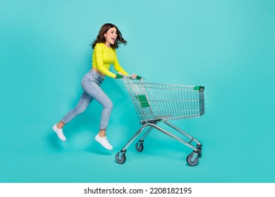 Full size profile photo of cool brunette hair young lady run with trolley wear shirt jeans sneakers isolated on teal color background