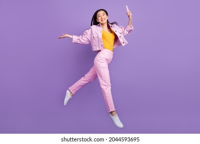 Full size profile photo of funky young brunette lady jump hold telephone wear jacket jeans sneakers isolated on violet background