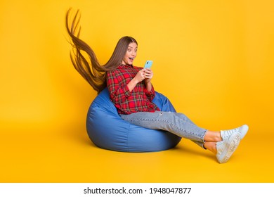 Full size profile photo of optimistic brunette long hairdo lady hold telephone wear shirt jeans sit on bag isolated on yellow background