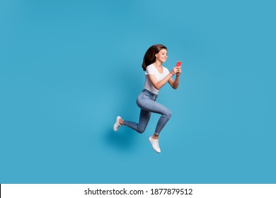 Full size profile side phot of brunette pretty girl run jump copyspace hold phone wear white t-shirt jeans isolated on blue color background