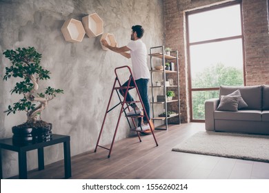 Full size profile view photo of mixed race guy installing wall shelf standing stepladder repair of new flat handmade working hanging last shelf checking sustainability flat indoors