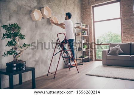 Full size profile view photo of mixed race guy installing wall shelf standing stepladder repair of new flat handmade working hanging last shelf checking sustainability flat indoors