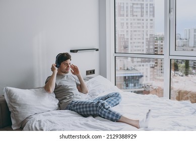 Full Size Satisfied Young Man Wear Grey T-shirt Blue Pajamas Headphones Sit In Bed Listen To Music Do Stretching Rest At Home On Weekends Relax Indoors Bedroom Good Mood Night Morning Bedtime Concept