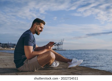 Full Size Side View Young Strong Sporty Fit Fun Sportsman Man In Sports Clothes Shorts Warm Up Train Using Mobile Cell Phone At Sunrise Sun Over Sea Beach Outdoor On Pier Seaside In Summer Day Morning