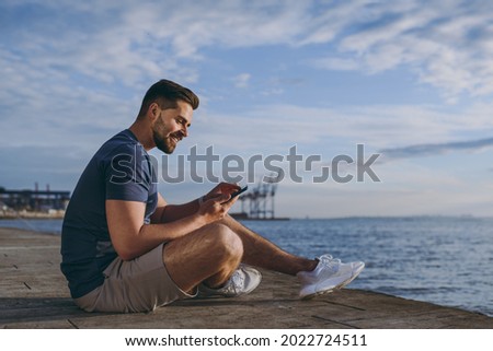 Full size side view young strong sporty fit fun sportsman man in sports clothes shorts warm up train using mobile cell phone at sunrise sun over sea beach outdoor on pier seaside in summer day morning
