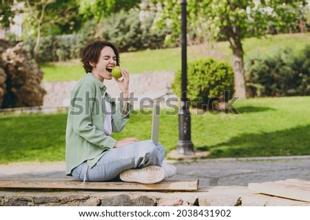 Full size side view young student freelancer woman 20s wearing green jacket jeans sit on bench in park outdoors use laptop pc computer biting apple fruit People vegetarian healthy lifestyle concept