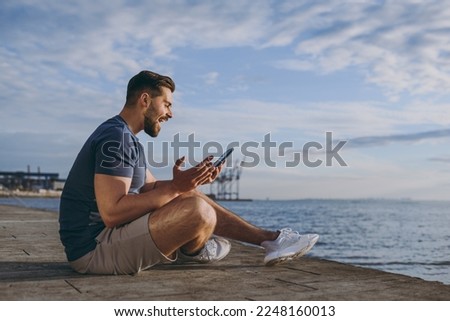 Full size side view young strong sporty fit sportsman man in sports clothes shorts warm up train chat by mobile cell phone at sunrise sun over sea beach outdoor on pier seaside in summer day morning