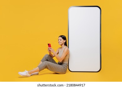 Chica latina feliz de tamaño completo y sonriente que lleva camisa de tanque sentada cerca de un gran teléfono móvil de pantalla en blanco con espacio de trabajo espacio de simulación usar smartphone aislado en el fondo amarillo plano