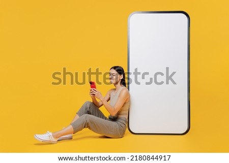 Full size smiling happy young latin woman she wear tank shirt sit near big huge blank screen mobile cell phone with workspace copy space mockup area use smartphone isolated on plain yellow backround