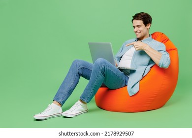 Full size smiling young brunet man 20s years old wears blue shirt sit in bag chair hold use work on laptop pc computer point index finger on screen isolated on plain green background studio portrait