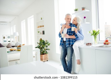 Full size of sweet stylish attractive couple, woman sitting on tabletop, man standing near her, looking at each other, having mug of tea in hands, enjoying time together in flat, apartment, indoor