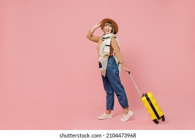 Full size traveler tourist mature elderly senior lady woman 55 years old wear brown shirt hat scarf hold suitcase bag go move look camera isolated on plain pastel light pink background studio portrait