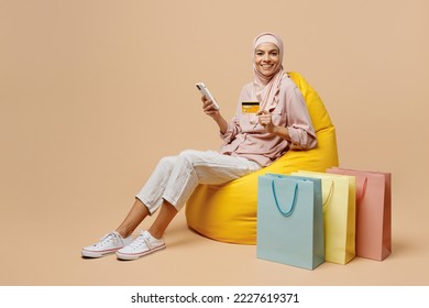 Full size young arabian asian muslim woman she wear abaya hijab sit in bag chair hold mobile phone credit card shopping isolated on plain light beige background. People uae islam religious concept. - Powered by Shutterstock