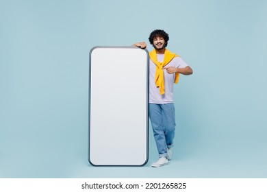 Full size young bearded Indian man 20s wears white t-shirt stand near pointing on big mobile cell phone with blank screen workspace area isolated on plain pastel light blue background studio portrait - Shutterstock ID 2201265825