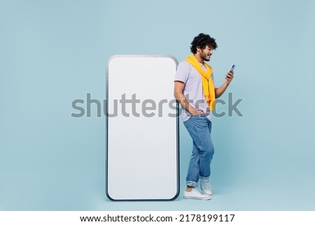 Full size young bearded Indian man 20s wears white t-shirt hold in hand stand near big mobile cell phone with blank screen workspace area isolated on plain pastel light blue background studio portrait