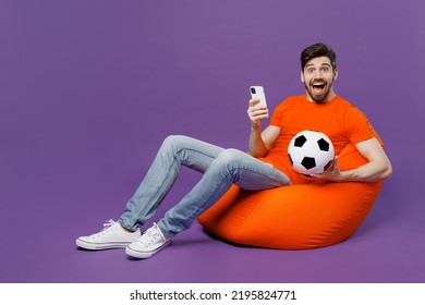 Full size young fan man he wear orange t-shirt cheer up support football sport team hold soccer ball mobile cell phone watch tv live stream sit in bag chair isolated on plain dark purple background - Shutterstock ID 2195824771