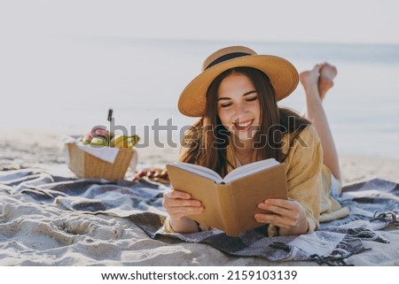 Full size young happy traveler tourist woman 20s in straw hat shirt summer clothes read book lying on plaid have picnic outdoor on sea sand beach background People vacation lifestyle journey concept