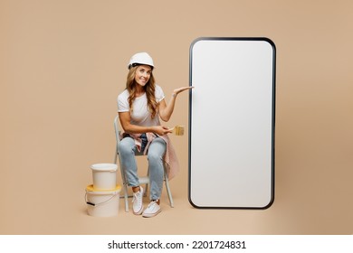 Full size young laborer handyman woman in white t-shirt helmet point blank screen area mobile cell phone hold brush paint isolated on plain beige background Instruments accessories for renovation room - Powered by Shutterstock