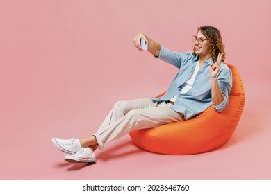 Full Size Young Man With Long Curly Hair In Blue Shirt Glasses Sit In Bag Chair Doing Selfie Shot On Mobile Cell Phone Post Photo Social Network Show V-sign Isolated On Pastel Plain Pink Background