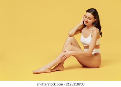 Full size young sexy brunette woman 20s wearing white underwear with perfect fit figure sitting on floor touch legs soft skin isolated on plain yellow color background. People female beauty concept.