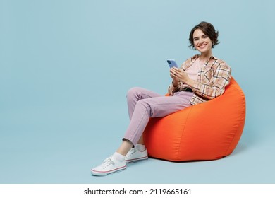Full size young smiling cheerful happy woman 20s in brown shirt sit in bag chair hold use mobile cell phone isolated on pastel plain light blue background studio portrait. People lifestyle concept.
