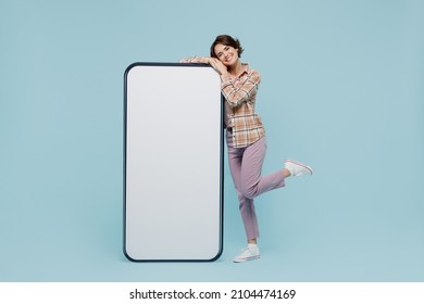 Full size young smiling happy woman 20s wear casual brown shirt stand near big mobile cell phone with blank screen workspace area isolated on pastel plain light blue color background studio portrait.