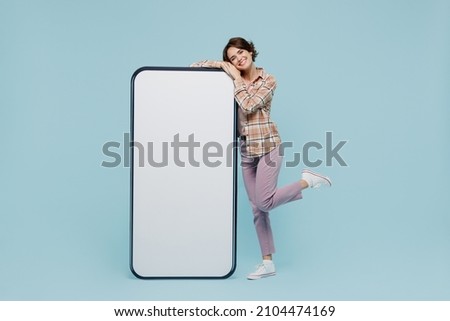 Full size young smiling happy woman 20s wear casual brown shirt stand near big mobile cell phone with blank screen workspace area isolated on pastel plain light blue color background studio portrait.