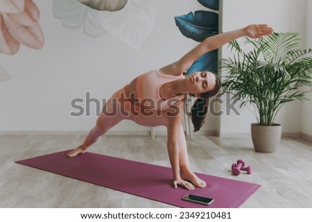 Full size young sporty athletic fitness trainer instructor woman in pink tracksuit doing yoga exercises asana near mobile cell phone stretch body with hands sit stretch on mat floor at home gym indoor