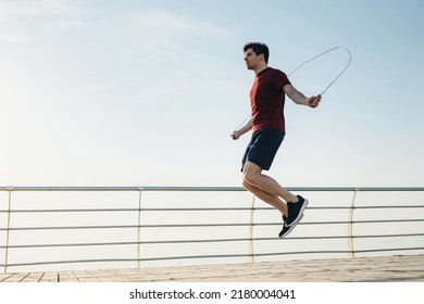 Full size young strong sporty toned fit sportsman man 20s wear sports clothes jumping on skipping rope warm up training at sunrise sun dawn over sea beach outdoor on pier seaside in summer day morning - Powered by Shutterstock