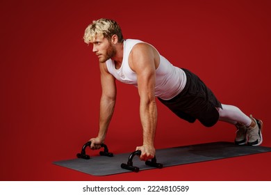 Full size young strong sporty sportsman man wear white clothes spend time in home gym train do push-ups exercises on floor with handles isolated on plain red background. Workout sport fit body concept - Powered by Shutterstock