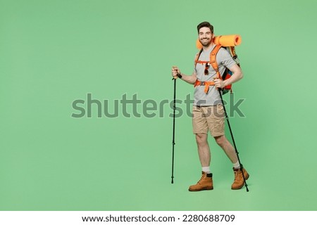 Full size young traveler white man carry backpack stuff mat walk with trakking poles isolated on plain green background. Tourist leads active healthy lifestyle. Hiking trek rest travel trip concept