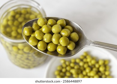 Full spoon of canned green peas close-up. Marinated sweet peas in a metal spoon over jar and plate. Snack, salad ingredient, vegetable protein, vegetarian food. Top view.