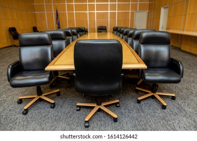 Full view of large black chair in conference room with other chairs facing away from it around light brown conference table Background has metal grid pattern wall with a round analog clock