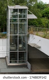 Full View Of A Transparent Glass Elevator Shaft That Provides An Insight Into The Drive Technology Of The Lift. The Elevator Is Located Outside A Tram Station Surrounded By Trees.