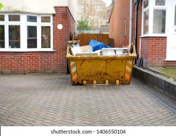 Full yellow rubbish skip on driveway, one side of frame. Space to add own text on pavement floor in front of the yellow bin & house. Great for background use, renovation, home clearance moving concept