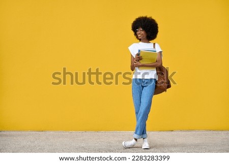 A fullbody portrait of african american girl with curly afro hairstyle stands with backpack and holds exercise books on a yellow background. Smart student looking aside on orange wall with copy space.