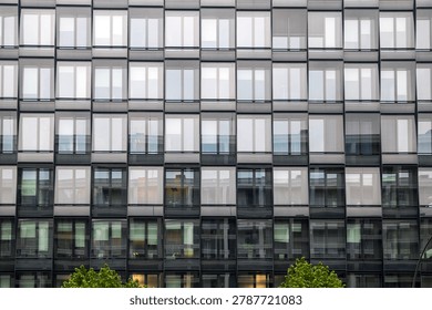 A full-frame architectural shot capturing the repeating geometric pattern of a modern glass office building facade. The facade consists of numerous rectangular window panes arranged in a strict grid, with alternating sections of white vertical blinds and dark glass panels. The lower windows clearly reflect the silhouette of another classical-style building across the street, creating a layered visual texture. The top of a green tree is visible at the very bottom edge.