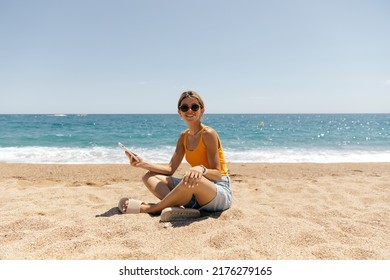 Full-lenght outdoor photo of smiling attractive woman with collected hair and tanned skin sitting on the beach with phone. Adorable girl using photo on the beach in sunlight - Powered by Shutterstock